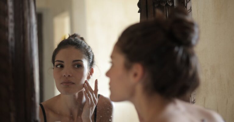 A woman checks her skin in the mirror, engaging in her morning skincare routine indoors.