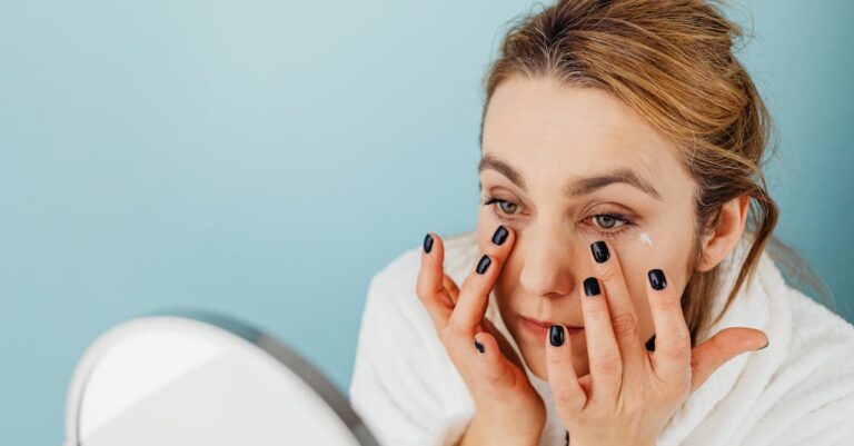Woman applies eye cream on her under-eye area with black nails, wearing a white robe.