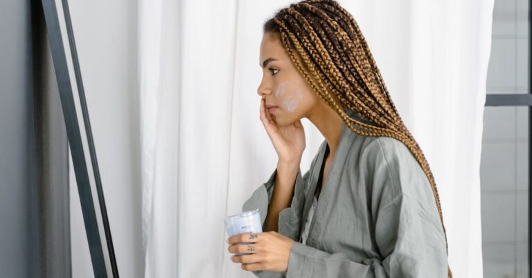 Young woman in robe applying moisturizer in front of mirror as part of her daily skincare routine.
