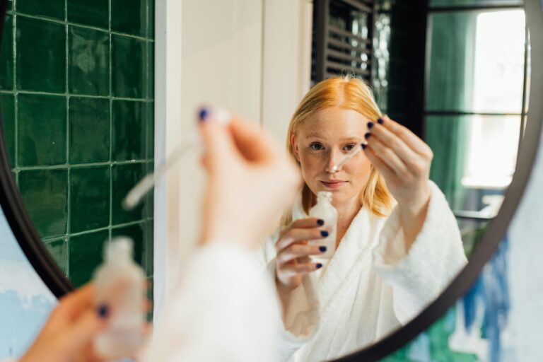 Adult woman applying skincare serum while wearing a bathrobe in front of a bathroom mirror.
