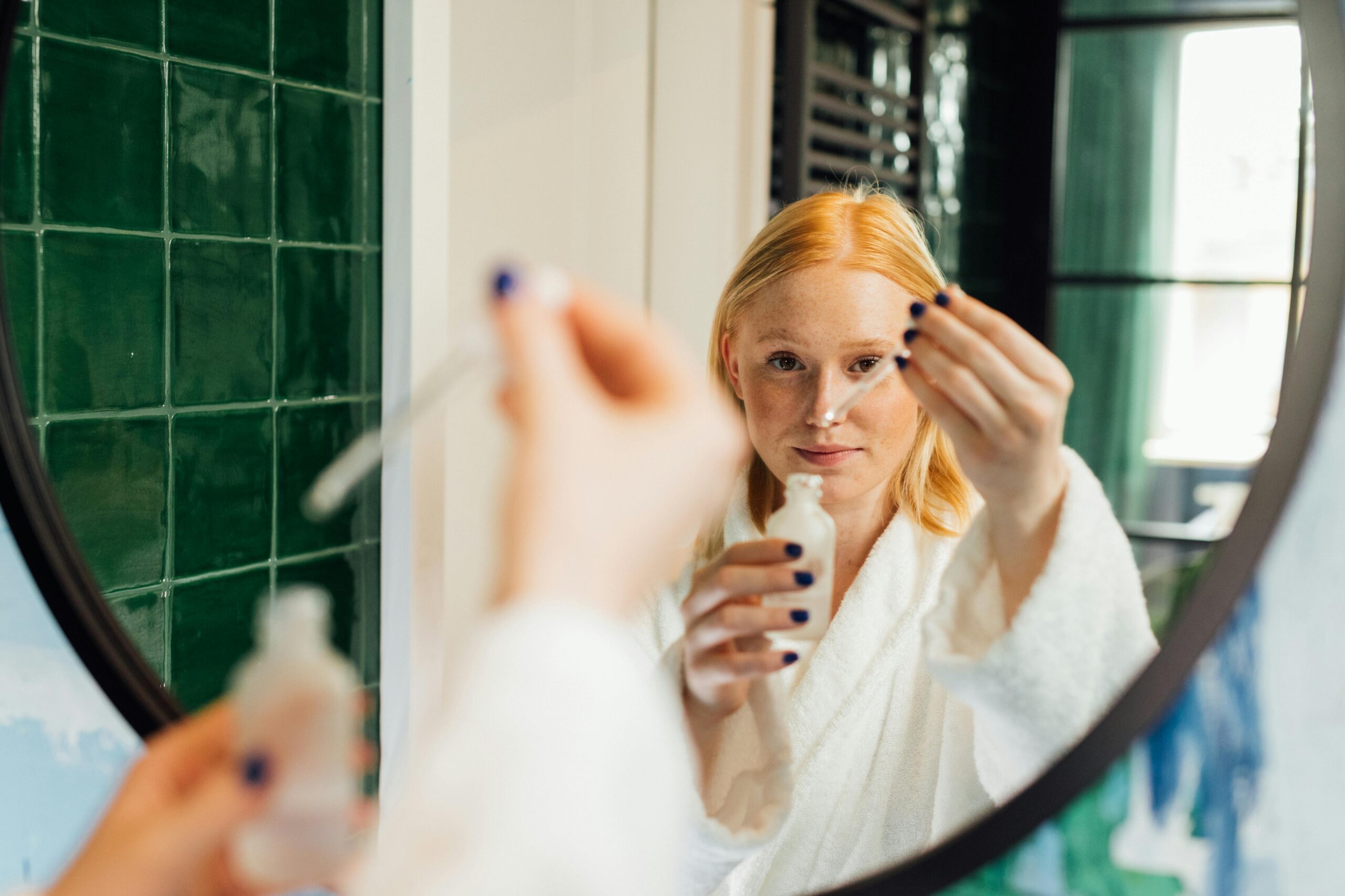 Adult woman applying skincare serum while wearing a bathrobe in front of a bathroom mirror.